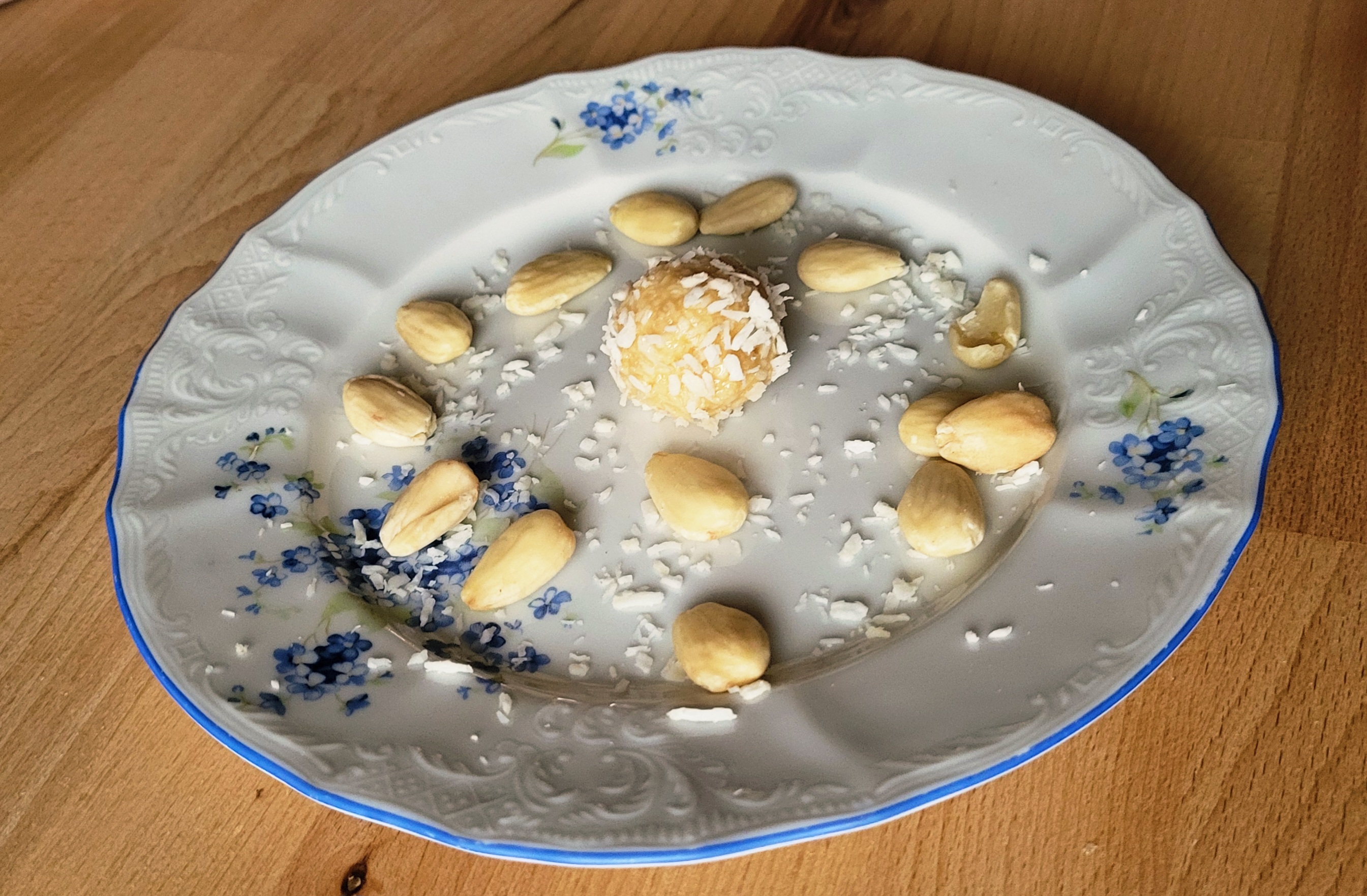 Nepecena kokosova kulicka obalena ve strouhanem kokosu na taliri na drevenem stole. Jako dekorace okolo strouhany kokos a mandle.